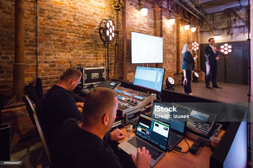 Business conference backstage Mixed sound board and computer screen on the foreground. Businessman and businessman during presentation standing on the stage in convention center. Performing Arts Event Stock Photo