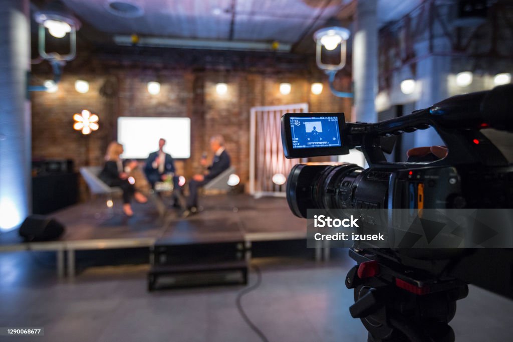 Business people during video conference Businesswoman and businessman giving presentation during online seminar, sitting on armchairs on the stage, holding microphones. Focus on video camera on the foreground. Home Video Camera Stock Photo