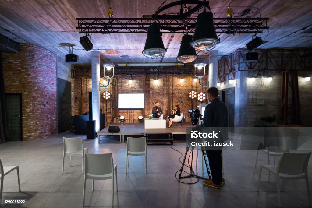 Business people during video conference Businesswoman and businessman giving presentation during online seminar, sitting on armchairs on the stage, holding microphones. Video camera on the foreground. Home Video Camera Stock Photo