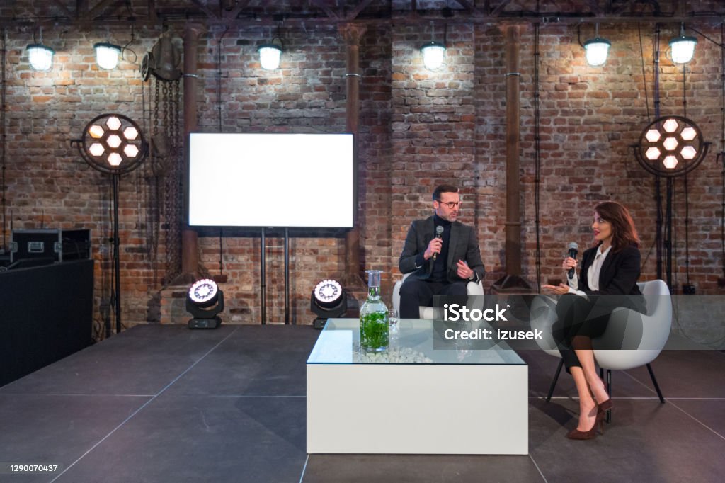 Businesswoman and businessman during presentation Businesswoman and businessman giving presentation during seminar, sitting on white armchairs on the stage, holding microphones. Stage - Performance Space Stock Photo