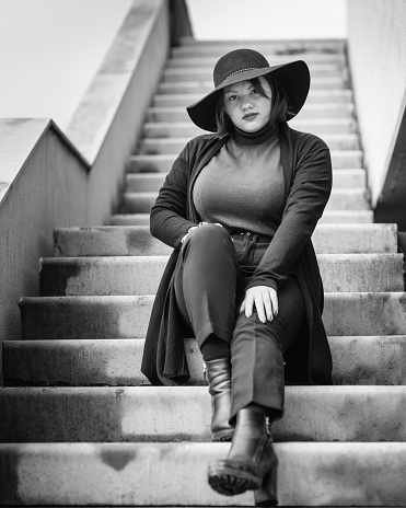 Brunette woman in hat sitting on the stairs of building in city. Plus size model. Black and white