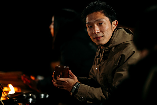 A portrait of a young man among a group of friends while sitting around a camp fire and enjoying food and drink at night in winter.