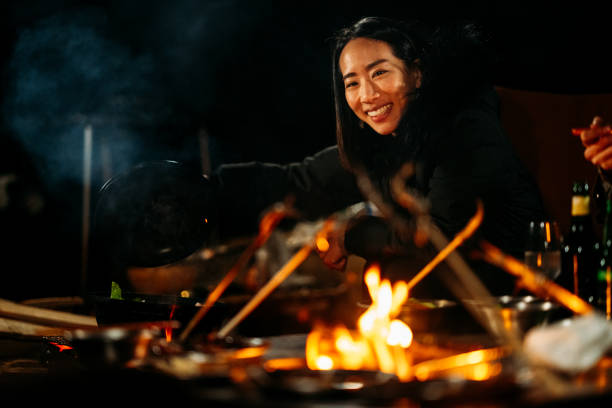 um retrato de uma jovem entre um grupo de amigos sentados ao redor do acampamento e apreciando comida e bebida à noite no inverno - bonfire people campfire men - fotografias e filmes do acervo