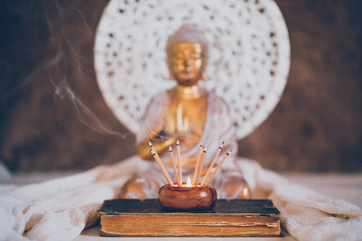 Fuming incense sticks on a book in front of a Buddha and a white mandala in the background. For mindfulness and wisdom concepts.  Selective focus and added grain. Part of a series.