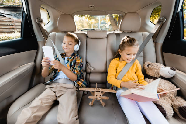 bambini sorridenti con smartphone e prenotare seduti vicino ai giocattoli sul sedile posteriore dell'auto - back seat foto e immagini stock