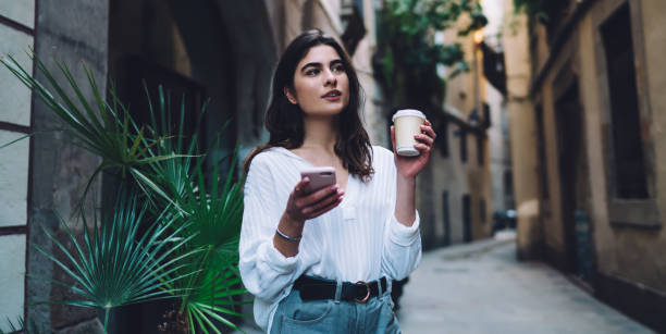 istock Contemplative hipster girl with disposable coffee cup thoughtful looking at architecture buildings during international travelling, Caucasian female generation Z with cellular device thinking outdoors 1290106537
