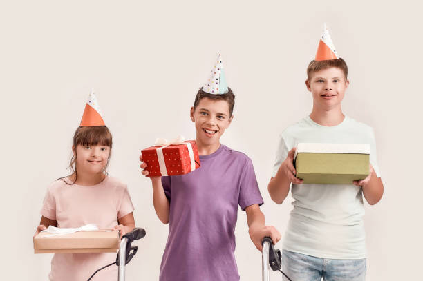trois enfants handicapés utilisant des chapeaux d’anniversaire souriant à l’appareil-photo et retenant des cadeaux tout en célébrant l’anniversaire ensemble d’isolement au-dessus du fond blanc - child cheerful little girls down syndrome photos et images de collection