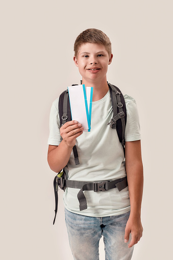 Happy disabled boy with Down syndrome wearing backpack smiling while holding air tickets, standing isolated over white background. Lifestyle of special children, Traveling concept. Vertical shot