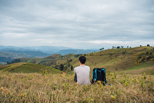Young asian man relaxing with camera bag on farmland hill in rural