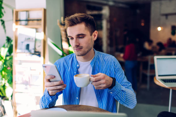 istock Handsome Caucasian man with cup of hot cappuccino using internet connection for application messaging, millennial male in trendy blue shirt browsing network website on modern cellphone gadget 1290146835