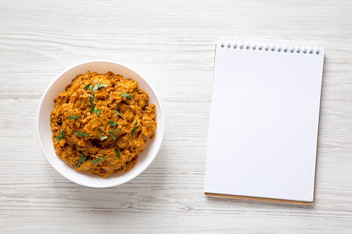 Homemade Chicken Tikka Masala in a white bowl, blank notepad on a white wooden background, top view. Flat lay, overhead, from above.