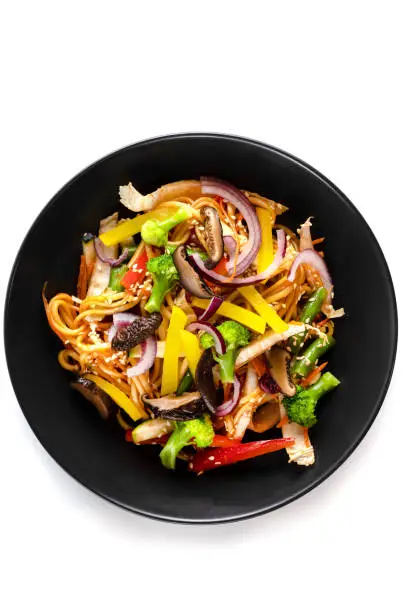 Photo of Sliced grilled vegetables, noodles in sauce, sesame seeds in a black plate isolated on white background, top view