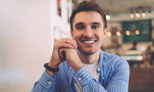 istock Half length portrait of cheerful Caucasian millennial keeping hands on cafeteria table during free time, prosperous male dressed in trendy blue shirt smiling at camera and posing in coffee shop 1290156310