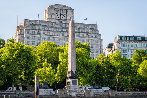 London, UK - June 22, 2018: Cleopatra's needle obelisk in city of Westminster by Ivybridge clock house on Thames river in summer green trees park with people