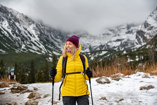 Blonde, smiling traveler woman in down jacket hiking with nordic walking poles on path in beautiful mountains.