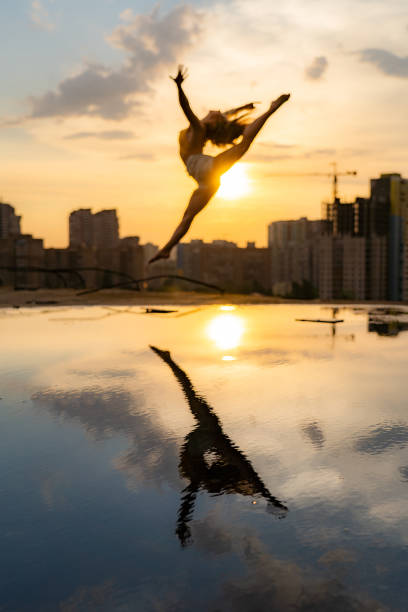 istock Female flexible dancer jumping during sunset on cityscape background with reflection in the water. Concept of freedom and happiness 1290167782