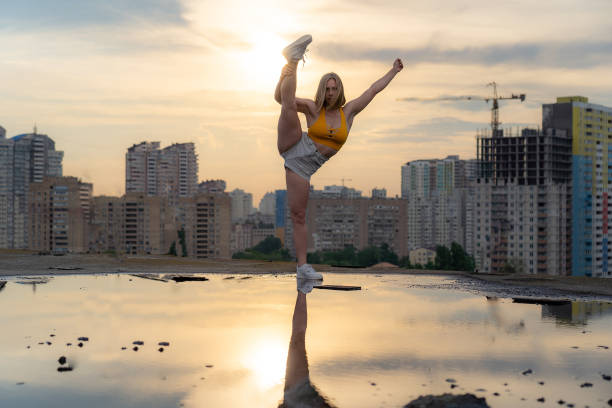 istock Flexible female gymnast standing split with reflection in the water during dramatic sunset with cityscape background 1290168118