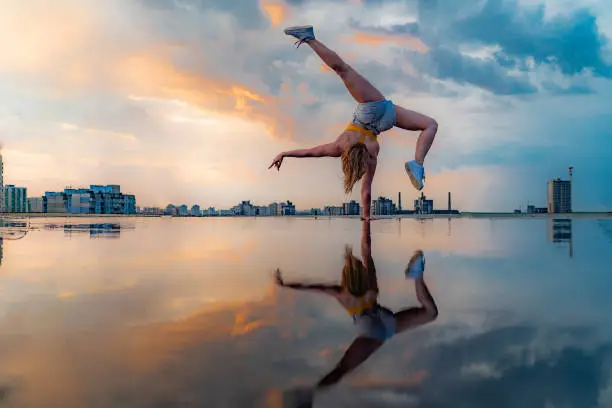 Female gymnast standing on one hand and keeping balance during dramatic sunset with reflection in the water of amazing clouds. Concept of Calisthenic, contortion and handstand.