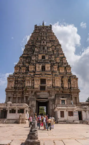 Hampi, Karnataka, India - November 4, 2013: Virupaksha Temple complex. Tall east Gopuram seen from courtyard inside said complex. Nandi statue and people under blue cloudscape.