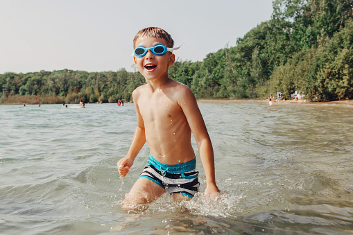 Cute funny Caucasian boy swimming in lake with underwater goggles. Child diving in water on beach. Authentic real lifestyle happy childhood. Summer fun outdoor seasonal activity.