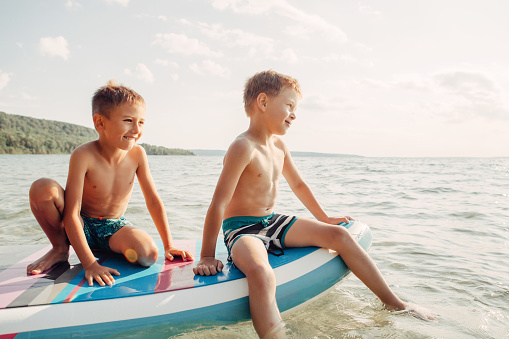 Two smiling Caucasian boys kids sitting on paddle sup surfboard in water. Children friends talking laughing. Modern outdoor summer activity. Aquatic recreation sport hobby. Happy childhood lifestyle.