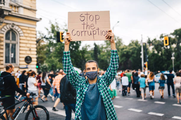 istock Young man with protective face mask, surrounded by a crowd, holding a poster in his hands. Young activist / protester against the corruption. Human rights and social issues concept. 1290189286