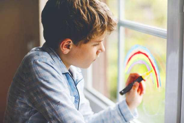 パンデミックコロナウイルス検疫中にカラフルな窓の色で虹を描くパジャマで素敵な学校の子供の男の子。世界中の虹を描く子供たちは、みんな元気になろう。 - epidemic paint virus illness ストックフォトと画像