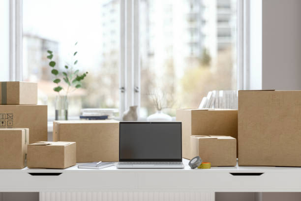 Working From Home According To New Normal With Laptop, Notepad, Adhesive Tape On The Table And Cardboard Boxes Prepared To Be Sent.