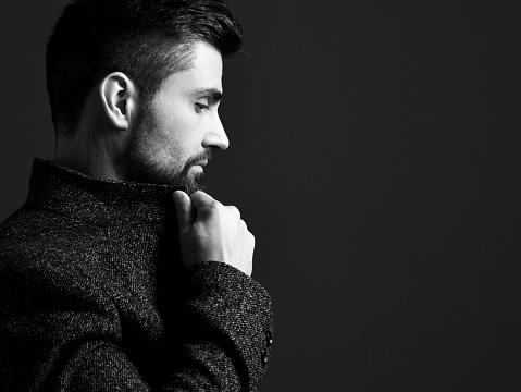 Black and white portrait in profile of serious bearded modern businessman in stylish tweed jacket holding his shirt collar over dark background. Stylish business casual style for men, portrait concept