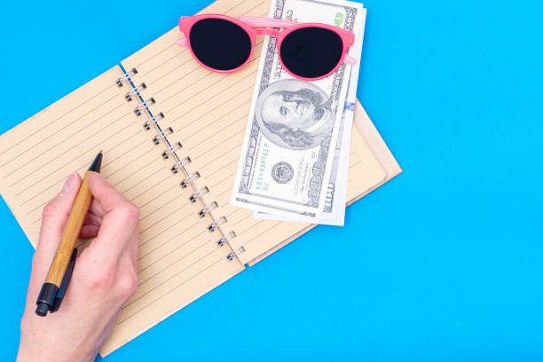 istock Travel planning, vacation concept. A hand holds a pen over an open blank notebook, dollars, pink sunglasses 1290212004