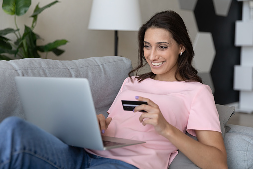 Happy young indian arab woman looking at computer screen, holding plastic bank credit card in hands, smiling female mixed race client involved in easy internet shopping, purchasing goods online.
