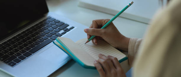 istock Female hand writing on blank notebook while working with laptop in office room 1290233024