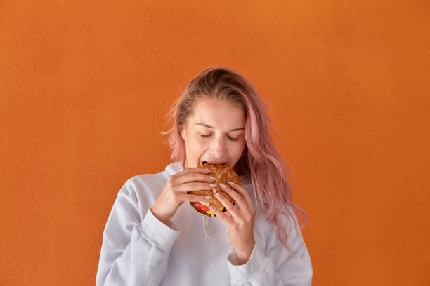 istock A woman on a diet eats a big Burger with an appetite. Orange background. 1290238646