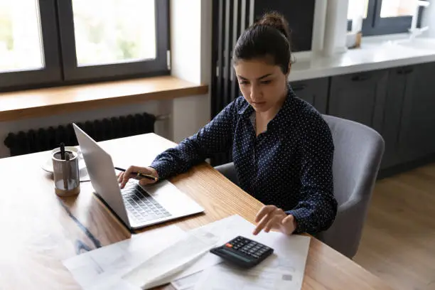 Bookkeeping. Busy millennial indian woman professional accountant preparing annual financial report checking result on calculator. Mixed race female freelancer counting tax rates declaring income