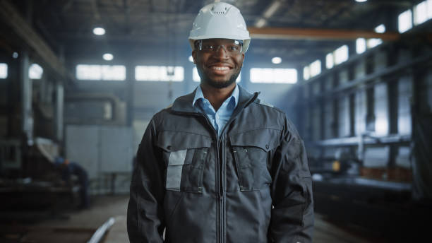 istock Happy Professional Heavy Industry Engineer/Worker Wearing Uniform, Glasses and Hard Hat in a Steel Factory. Smiling African American Industrial Specialist Standing in a Metal Construction Manufacture. 1290244364