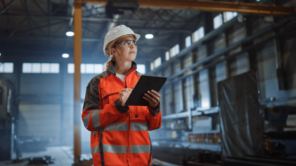 Professional Heavy Industry Engineer Worker Wearing Safety Uniform and Hard Hat, Using Tablet Computer. Serious Successful Female Industrial Specialist Walking in a Metal Manufacture Warehouse. profesional ingeniero de la industria pesada trabajador que lleva uniforme de seguridad y sombrero duro, utilizando la computadora de la tableta. seria especialista industrial femenina exitosa caminando en un almacén de fabricación de metales.