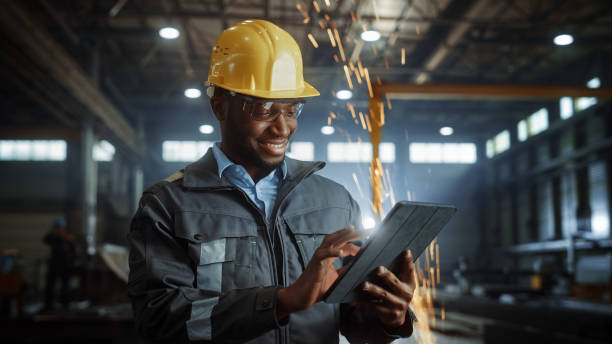 istock Professional Heavy Industry Engineer/Worker Wearing Safety Uniform and Hard Hat Uses Tablet Computer. Smiling African American Industrial Specialist Standing in a Metal Construction Manufacture. 1290244395