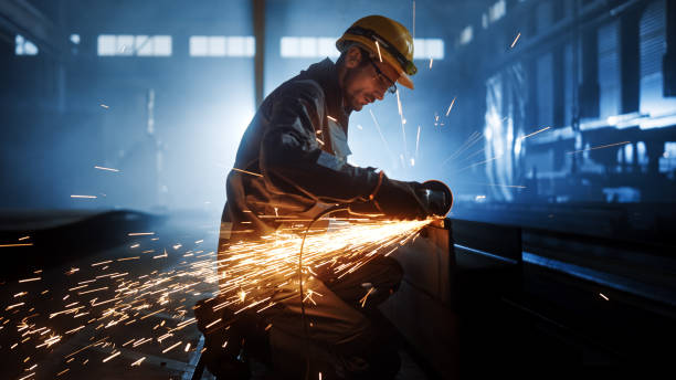 istock Heavy Industry Engineering Factory Interior with Industrial Worker Using Angle Grinder and Cutting a Metal Tube. Contractor in Safety Uniform and Hard Hat Manufacturing Metal Structures. 1290244404