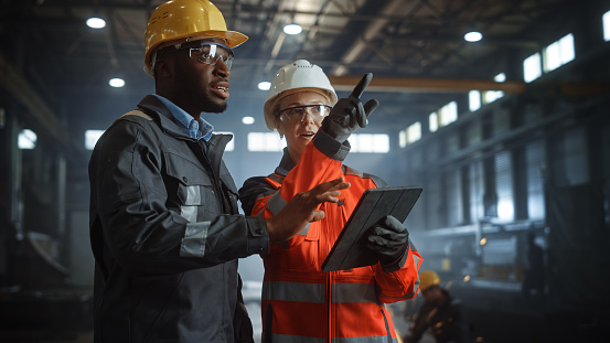 Dos ingenieros de la industria pesada están en la fábrica de fabricación de metales de acero, utilizan la tableta digital y tienen una discusión. Negro Afroamericano Especialista Industrial Hablar con la Técnica Femenina. photo