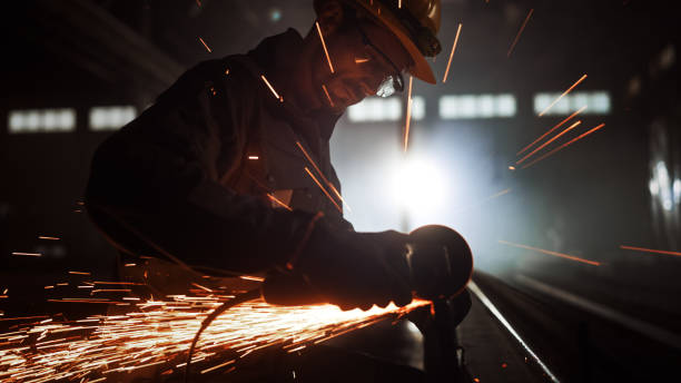 istock Heavy Industry Engineering Factory Interior with Industrial Worker Using Angle Grinder and Cutting a Metal Tube. Contractor in Safety Uniform and Hard Hat Manufacturing Metal Structures. 1290244411