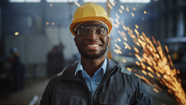 feliz profissional engenheiro da indústria pesada/trabalhador vestindo uniforme, óculos e chapéu duro em uma fábrica de aço. sorridente especialista industrial afro-americano em pé em uma fabricação de construção de metal. - work wear factory people occupation - fotografias e filmes do acervo