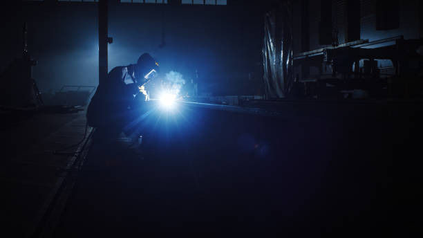 istock Heavy Industry Engineering Factory Interior with Industrial Worker Using a Welding Machine and Working on a Metal Tube. Contractor in Safety Uniform and Hard Hat Manufacturing Metal Structures. 1290244416