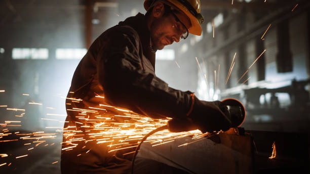 istock Heavy Industry Engineering Factory Interior with Industrial Worker Using Angle Grinder and Cutting a Metal Tube. Contractor in Safety Uniform and Hard Hat Manufacturing Metal Structures. 1290244421