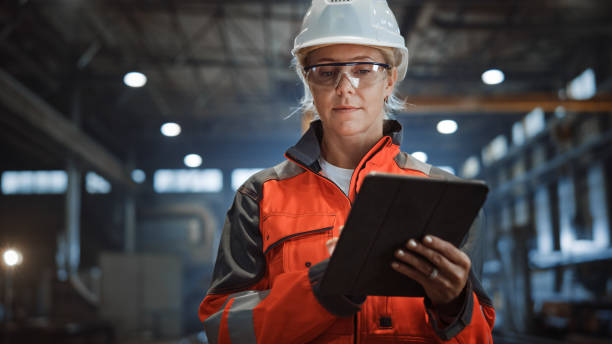 안전 유니폼과 하드 햇을 착용한 전문 중공업 엔지니어/작업자는 태블릿 컴퓨터를 사용합니다. 금속 제조 창고에서 걷는 심각한 성공적인 여성 산업 전문가. - construction safety protective workwear hardhat 뉴스 사진 이미지