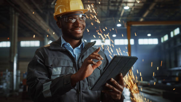 istock Professional Heavy Industry Engineer/Worker Wearing Safety Uniform and Hard Hat Uses Tablet Computer. Smiling African American Industrial Specialist Standing in a Metal Construction Manufacture. 1290244433