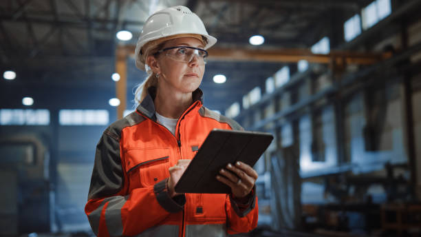 istock Professional Heavy Industry Engineer/Worker Wearing Safety Uniform and Hard Hat Uses Tablet Computer. Serious Successful Female Industrial Specialist Walking in a Metal Manufacture Warehouse. 1290244436