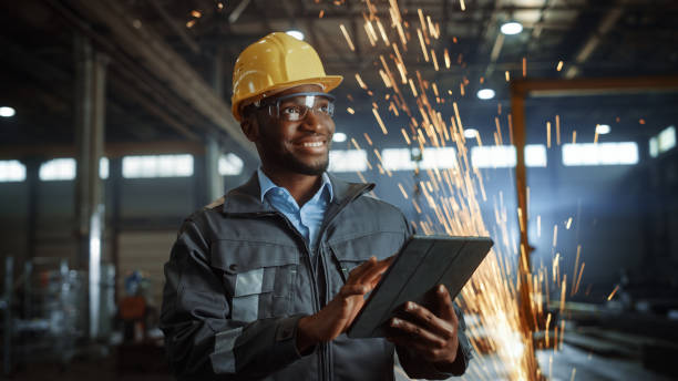 istock Professional Heavy Industry Engineer/Worker Wearing Safety Uniform and Hard Hat Uses Tablet Computer. Smiling African American Industrial Specialist Standing in a Metal Construction Manufacture. 1290244437
