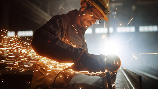 Heavy Industry Engineering Factory Interior with Industrial Worker Using Angle Grinder and Cutting a Metal Tube. Contractor in Safety Uniform and Hard Hat Manufacturing Metal Structures.