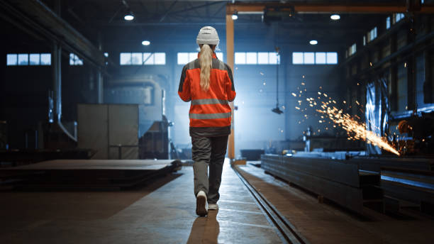 istock Professional Heavy Industry Engineer/Worker Wearing Safety Uniform and Hard Hat Uses Tablet Computer. Serious Successful Female Industrial Specialist Walking in a Metal Manufacture Warehouse. 1290244453