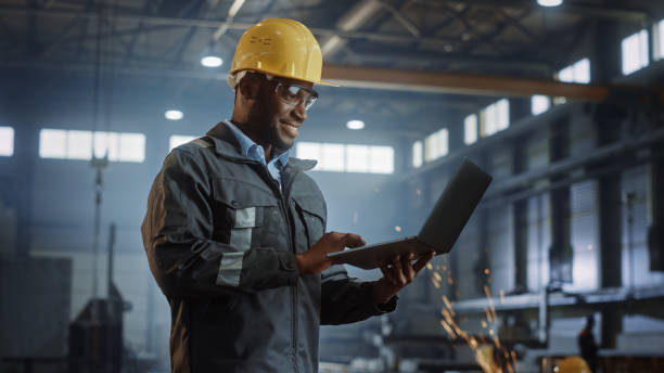 istock Professional Heavy Industry Engineer/Worker Wearing Safety Uniform and Hard Hat Uses Laptop Computer. Smiling African American Industrial Specialist Standing in a Metal Construction Manufacture. 1290244464
